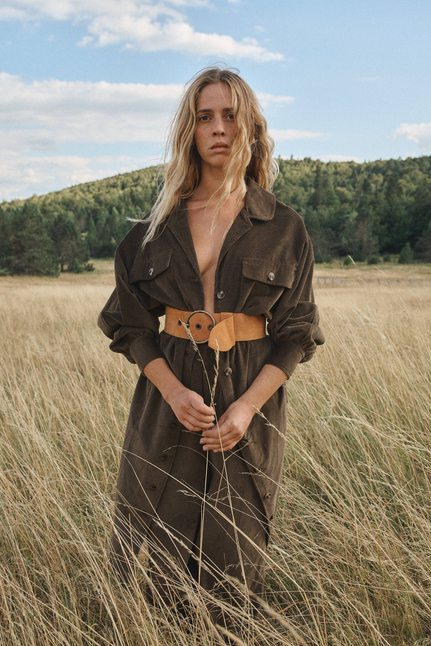 Femme debout dans un champ d’herbes sèches, portant une robe longue marron avec une ceinture cognac à boucle ronde, sous un ciel partiellement nuageux.