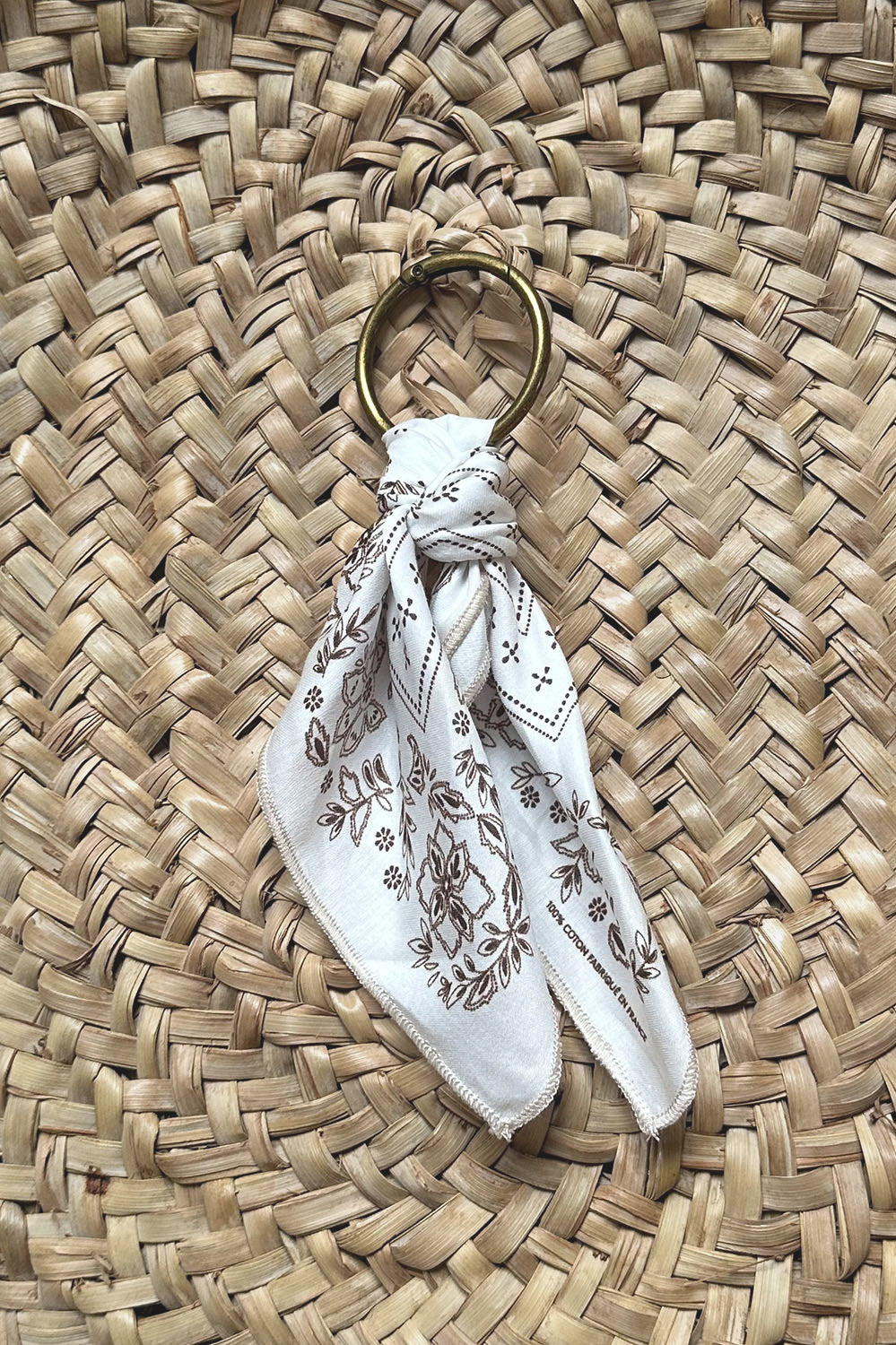 Porte-clés élégant avec foulard en coton blanc à motifs floraux marron, posé sur un tissage naturel en osier, alliant style bohème et savoir-faire artisanal.