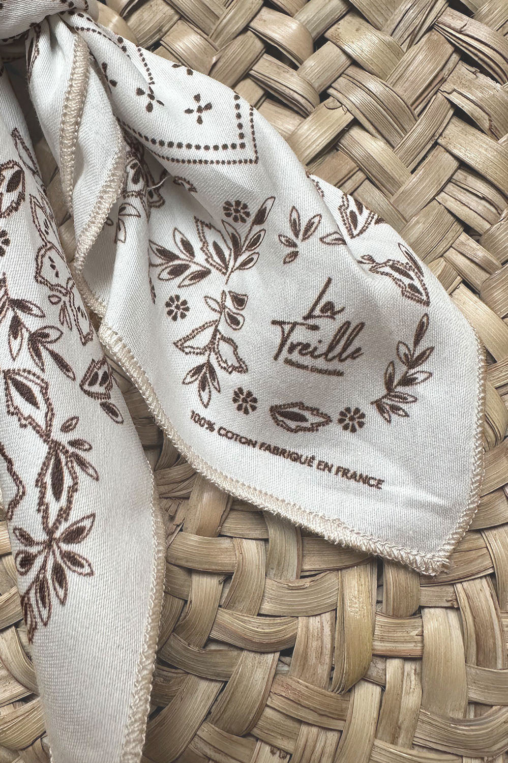 Carré en coton blanc orné de motifs floraux bruns et d’inscriptions indiquant une fabrication française, posé sur une surface tressée en osier de style bohème.