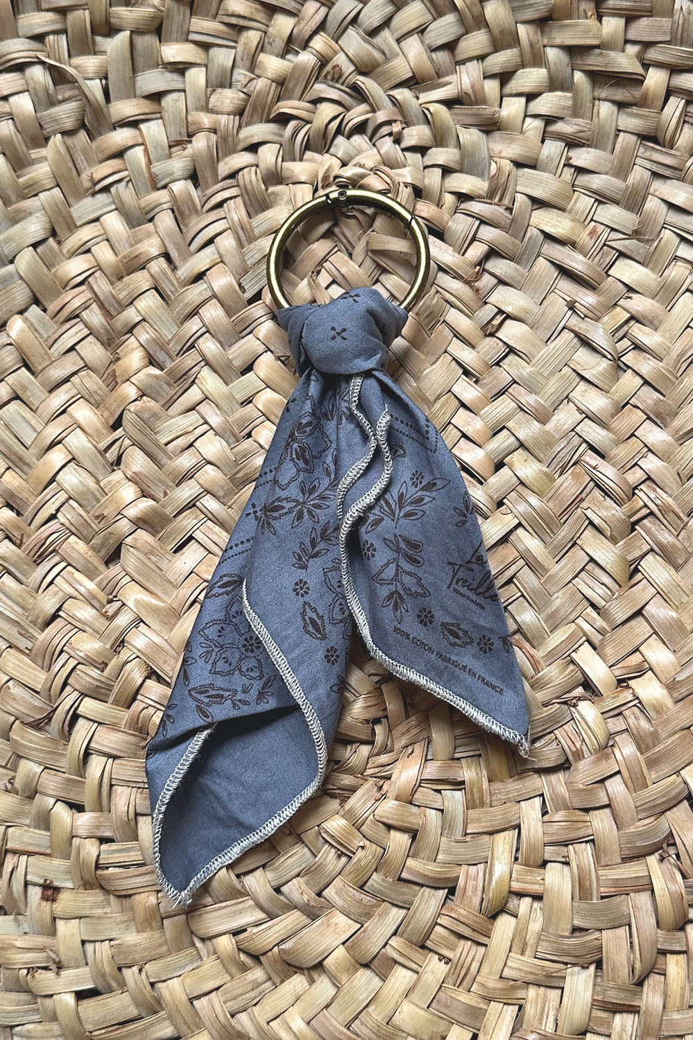 Porte-clés avec petit ours en tissu bleu denim imprimé floral, suspendu sur un fond en osier naturel, illustrant un savoir-faire artisanal et une finition soignée.
