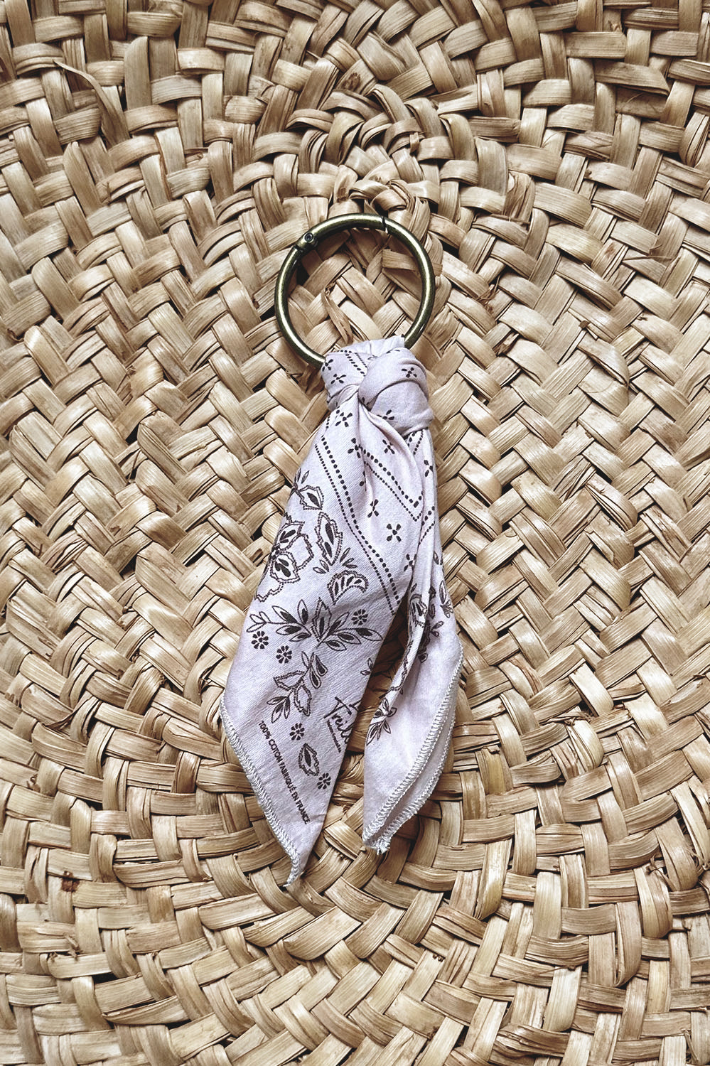 Porte-clés rond en métal doré avec un foulard en coton de couleur pétale orné de motifs floraux délicats, posé sur un fond en osier tressé naturel.