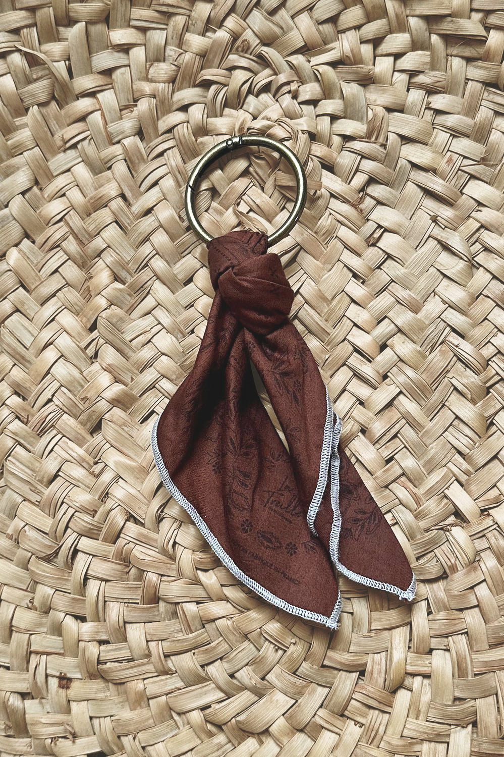Anneau doré avec foulard en coton couleur rouille à motifs subtils, présenté sur un fond en tissage naturel, évoquant un savoir-faire artisanal et une touche bohème.