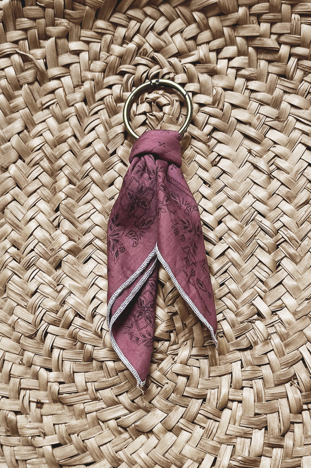 Porte-clés circulaire en métal doré orné d'un tissu en coton couleur sangria à motifs floraux subtils, posé sur une surface en osier tressé naturel.