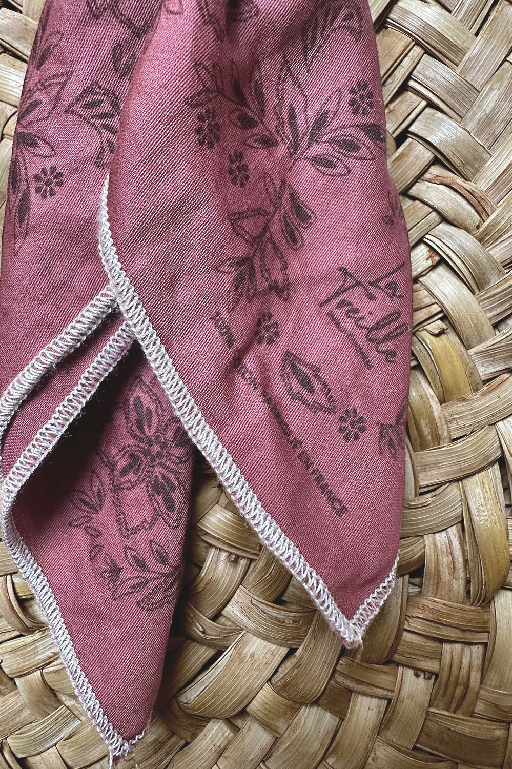 Tissu en coton teint sangria avec motifs floraux imprimés et finitions délicates, posé sur un panier tressé en osier, évoquant un style bohème et artisanal.