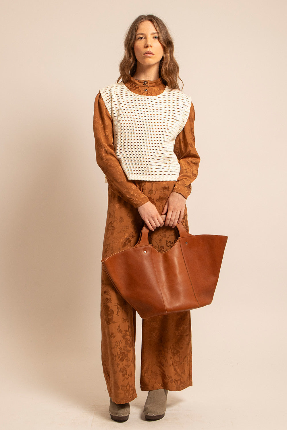 Femme debout portant un ensemble marron aux motifs subtils, un gilet crocheté blanc et tenant un grand cabas en cuir cognac fait main, évoquant un style bohème et élégant.