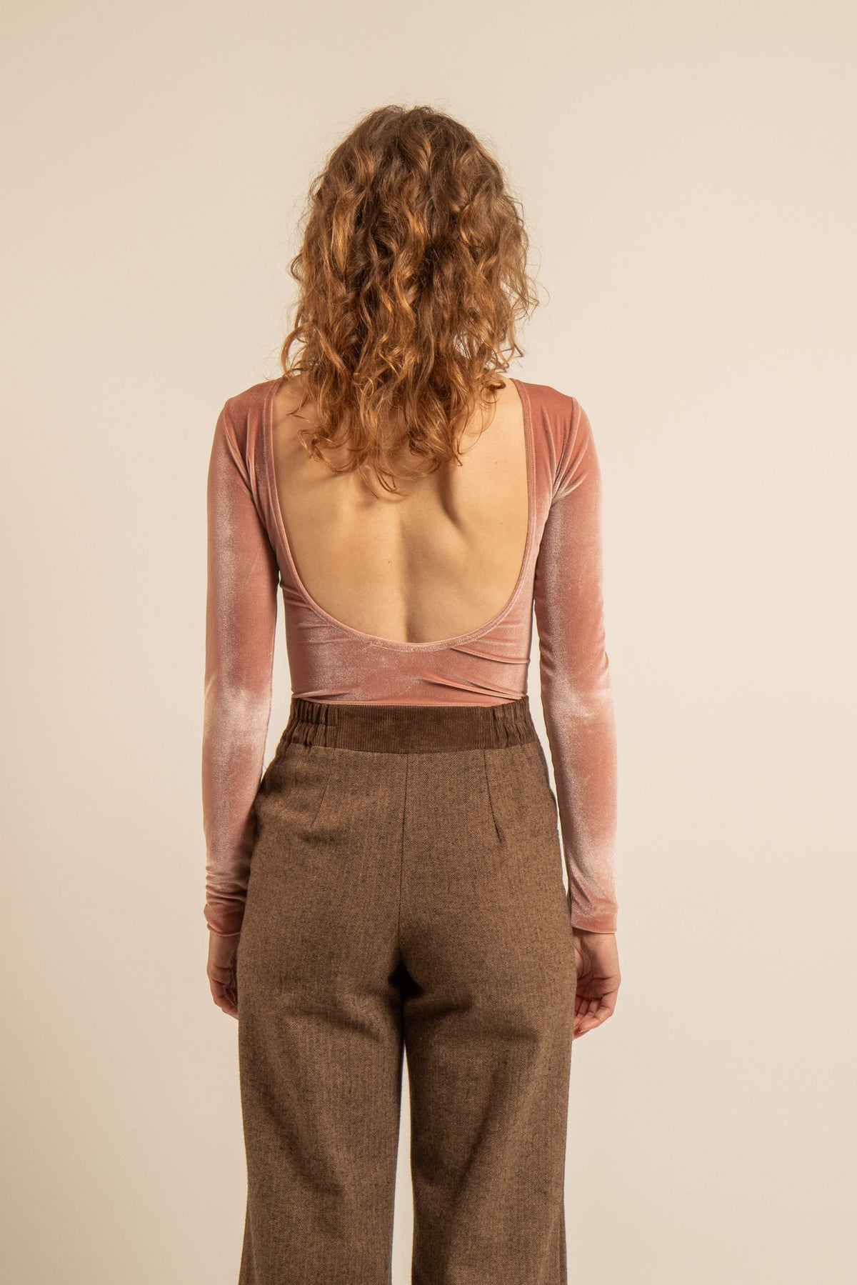 Vue de dos d’un body Cherry en velours rose poudré avec dos nu et manches longues porté avec un pantalon brun.