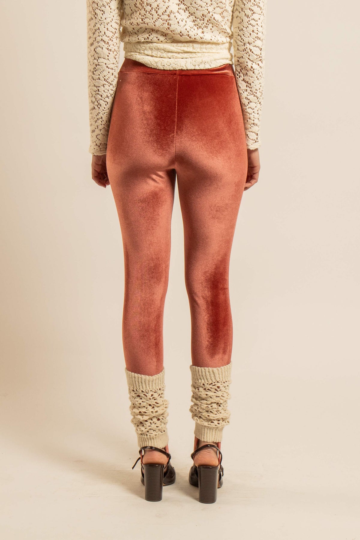 Vue arrière d’une femme portant des guêtres crème en crochet fait main sur un pantalon près du corps couleur terracotta, avec des escarpins noirs.