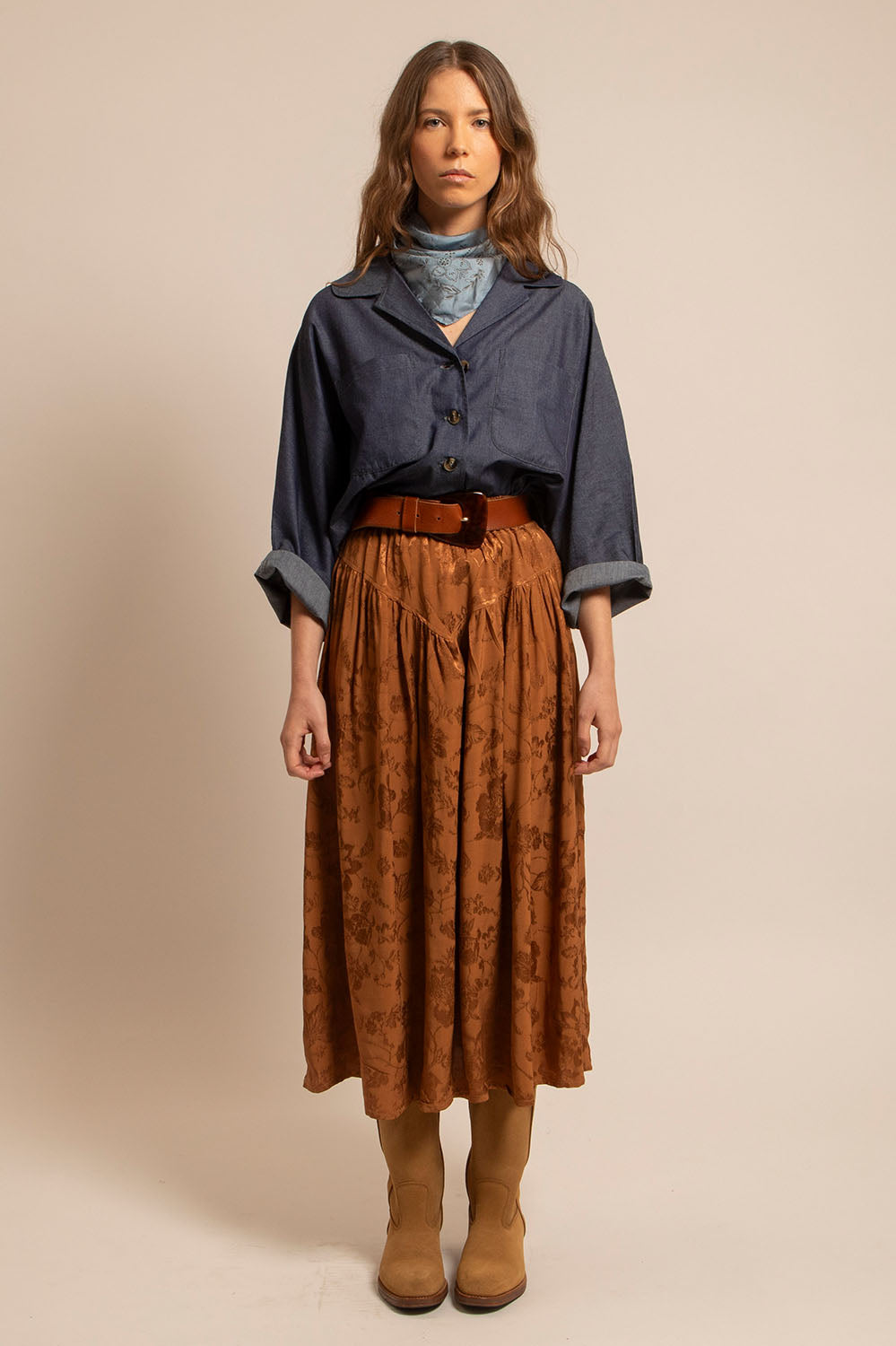 Femme portant la chemise Romy en chambray bleu brut, associée à une jupe fluide camel et des bottes beiges.