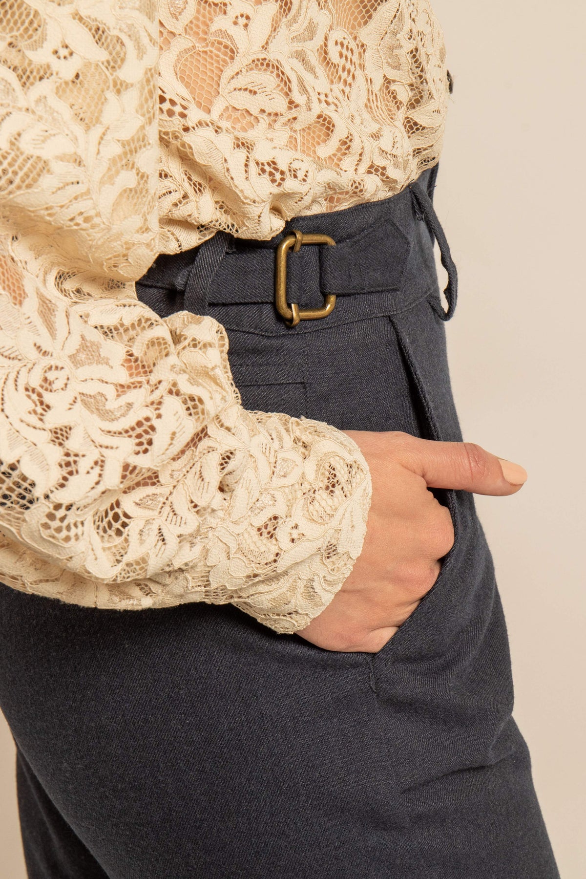 Pantalon bleu nuit en drap de laine avec martingale en laiton et poche latérale porté avec un chemisier en dentelle écrue.