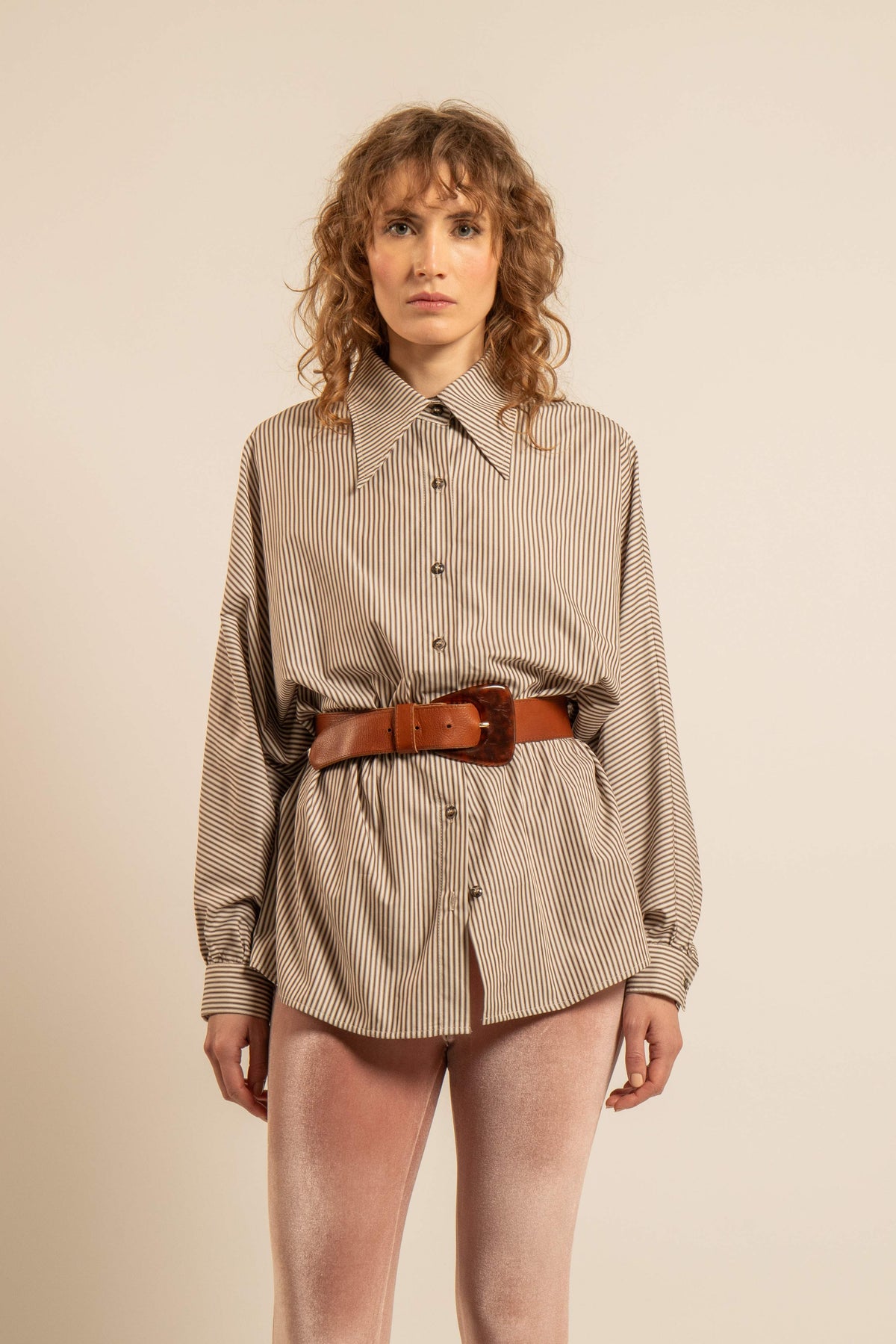 Femme portant une chemise oversize à fines rayures beige et marron avec col pointu manches chauve souris et ceinture en cuir brun devant un fond beige
