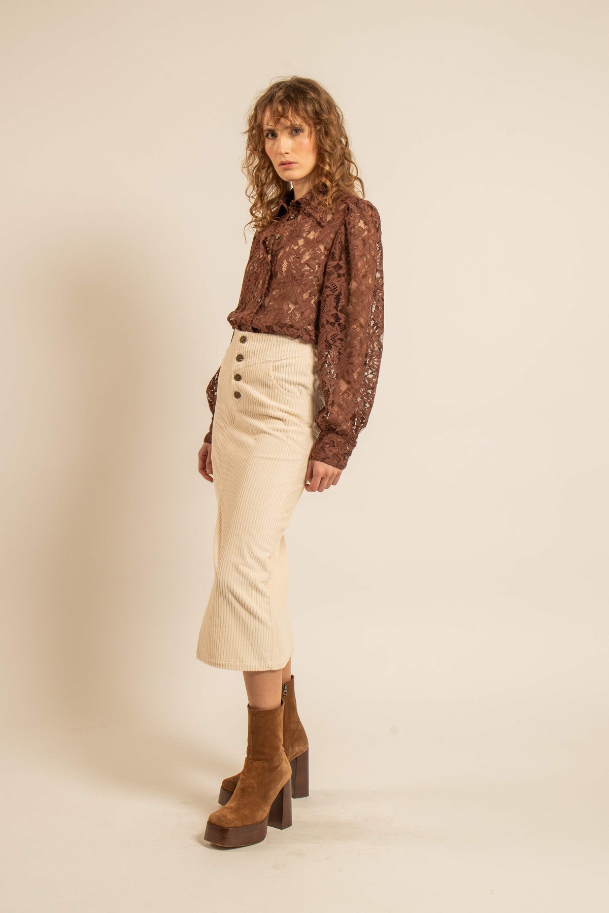 Modèle portant une jupe droite crème en velours côtelé avec taille haute et boutons devant accompagnée d’une blouse en dentelle brune et de bottines à talons