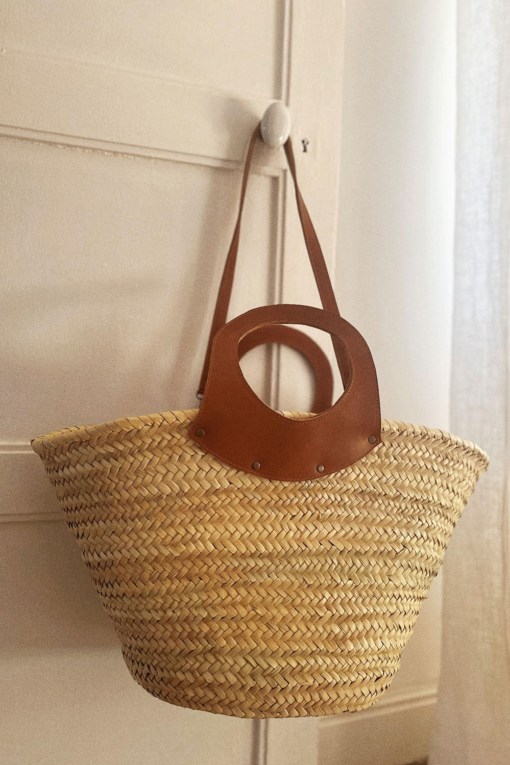 Panier en osier avec sangles longues en cuir pour un porté épaule, suspendu à une porte claire