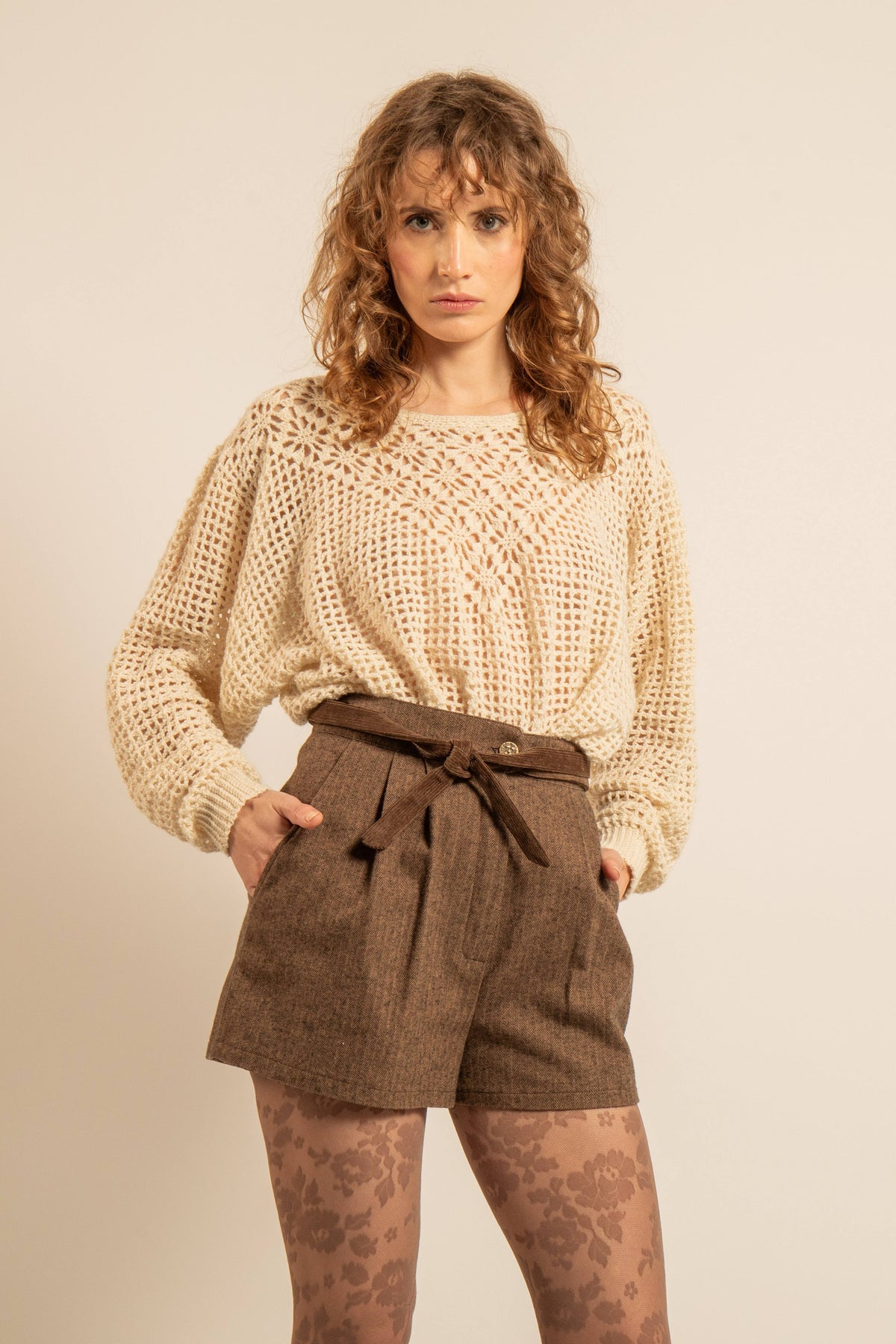 Short marron taille haute ceinturé porté avec un pull ajouré écru et des collants à motifs floraux dans un style vintage et décontracté
