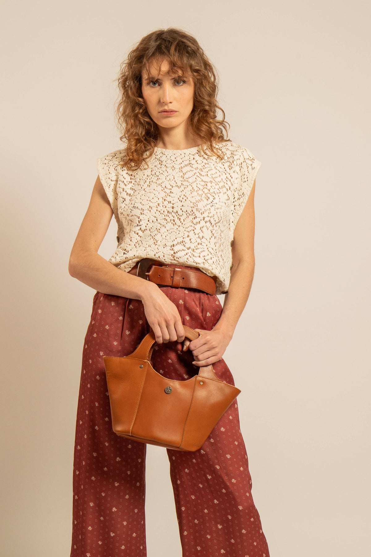 Femme portant un top en crochet ajouré écru sans manches, associé à un pantalon rouge imprimé, une ceinture marron et un sac en cuir camel.