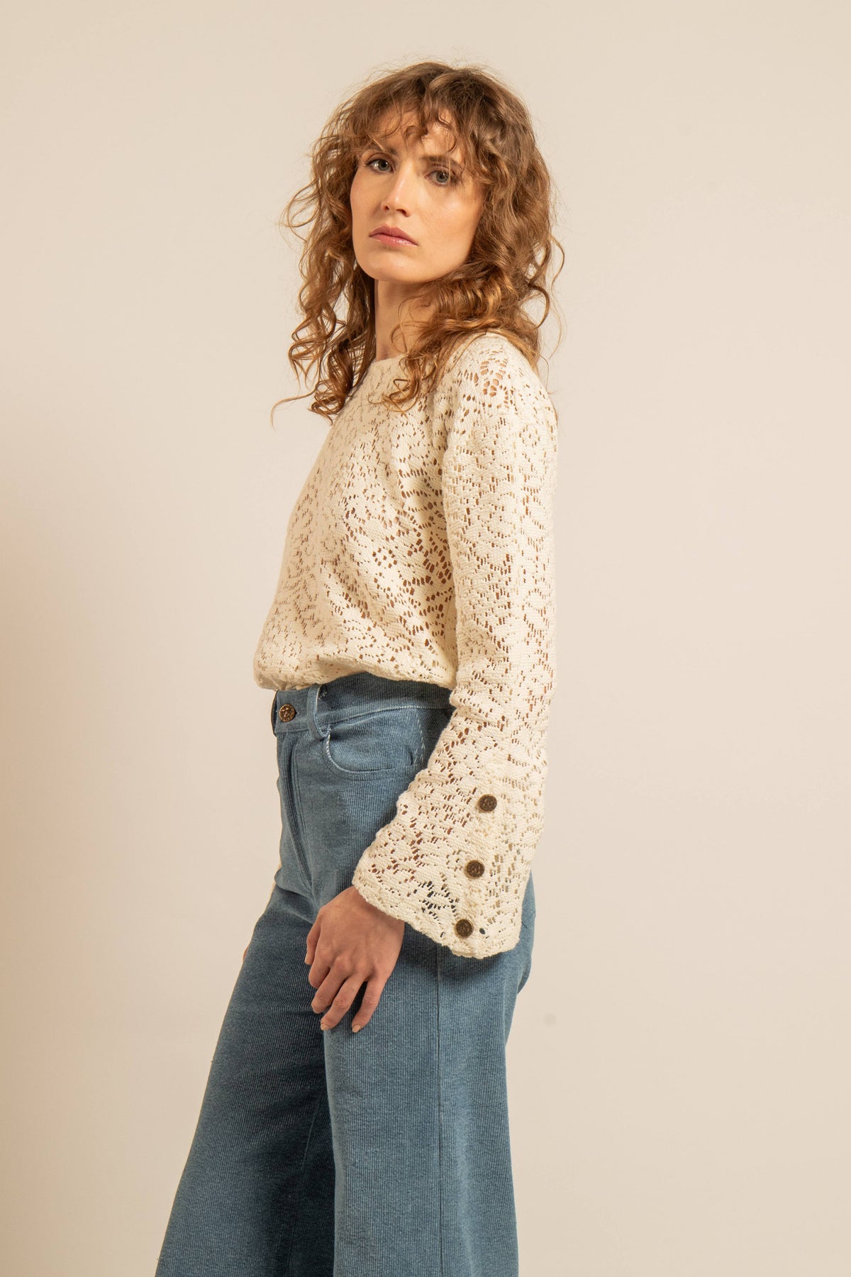 Top en maille crochet ajourée couleur naturel avec manches évasées et boutons Soleil en laiton aux poignets porté avec un jean par une femme pose de profil