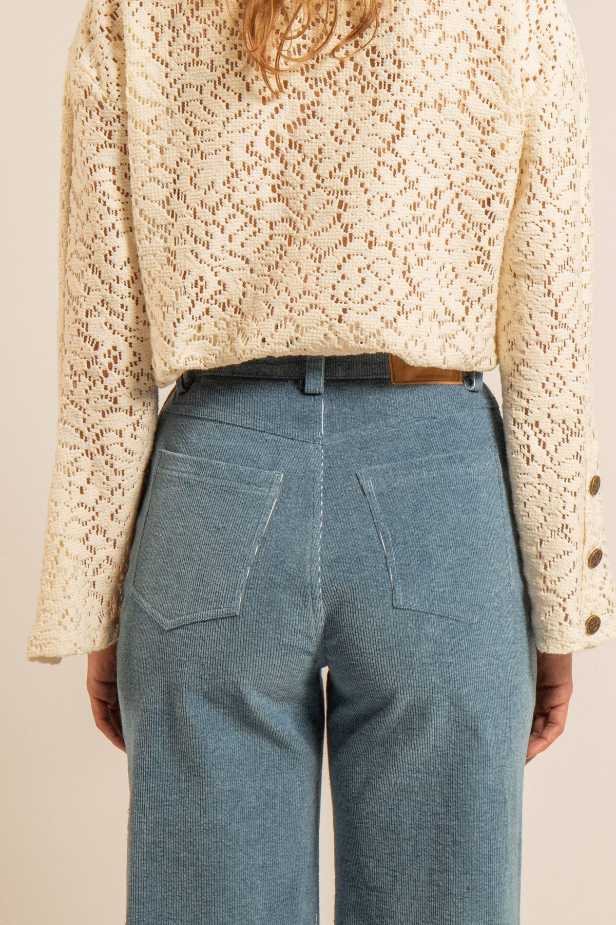 Top en maille crochet écrue légèrement transparente, manches évasées et boutons en laiton aux poignets, porté avec un pantalon bleu taille haute.