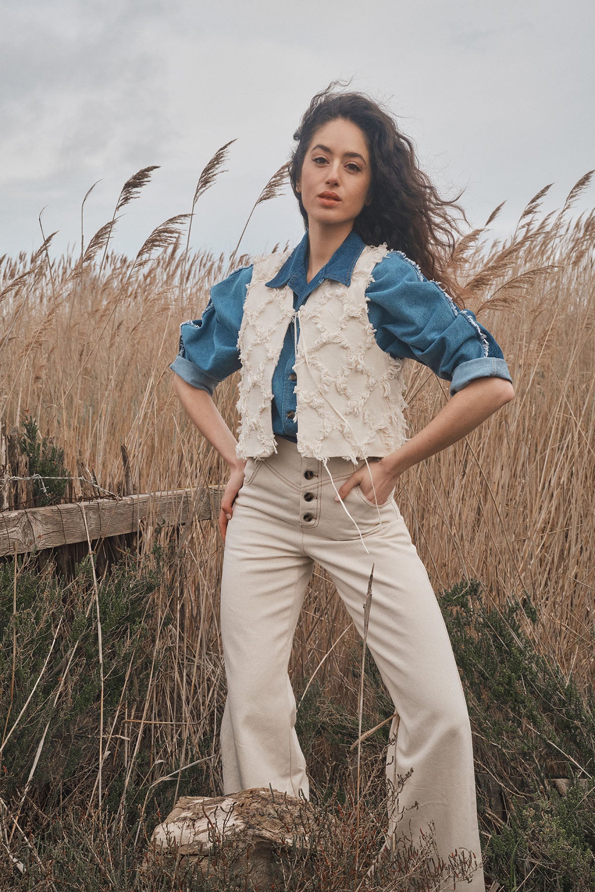 Femme debout dans un champ de hautes herbes, portant une chemise bleue, un gilet en crochet beige et un pantalon taille haute clair, avec une expression confiante.
