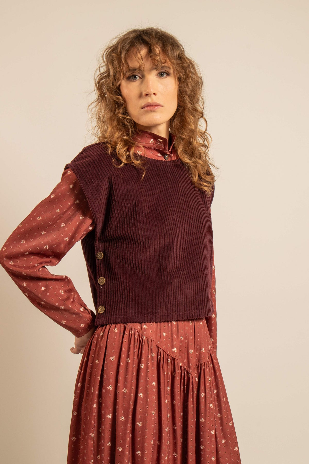 Top bordeaux en velours côtelé, sans manches et boutonné sur les côtés, porté sur une blouse imprimée à manches longues pour un look automnal.