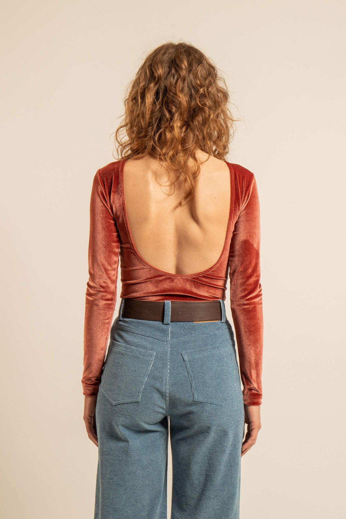 Vue de dos d’une femme portant un body en velours rouge à dos nu et manches longues avec un jean taille haute bleu.