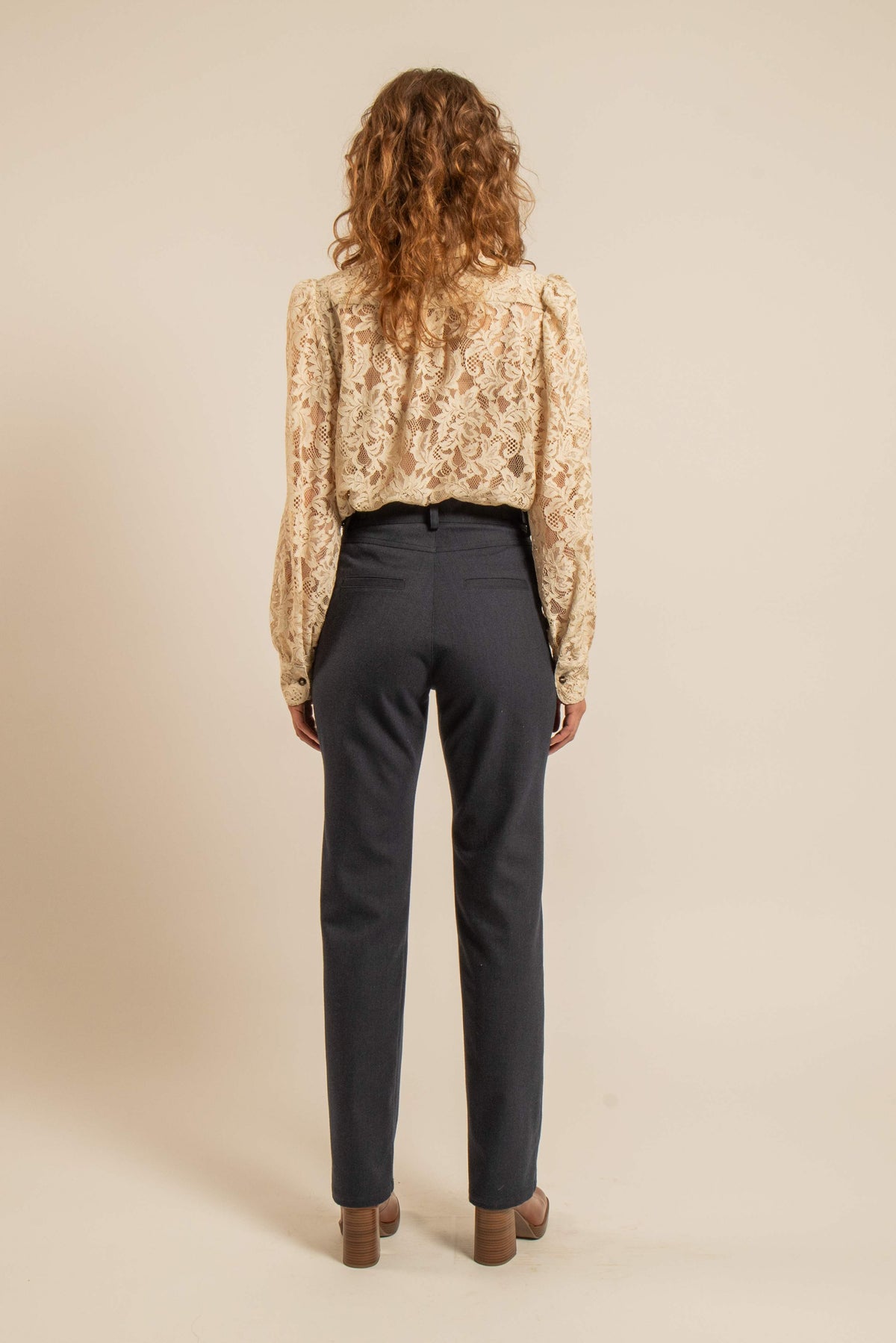 Vue de dos d’une femme portant une chemise en dentelle beige clair à manches bouffantes avec un pantalon noir dans un style inspiré des années 70