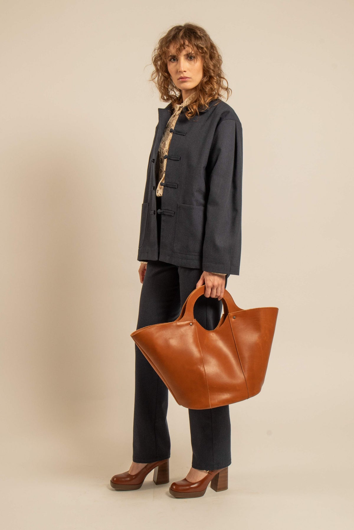 Femme portant une veste ample en laine bleu nuit à boutons tressés, avec pantalon sombre, escarpins bruns et grand sac cabas en cuir fauve.