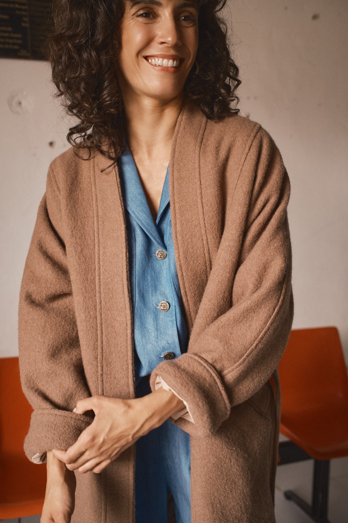 Femme souriante portant un manteau long oversize camel en laine à coupe kimono sur une chemise bleue dans un intérieur chaleureux
